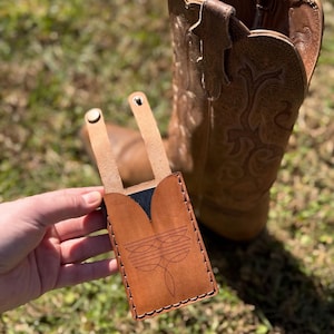May include: A brown leather phone case with a decorative stitched design, held in front of a brown cowboy boot. The case has a flap with two rivets and a stitched border. The background is blurred green grass.