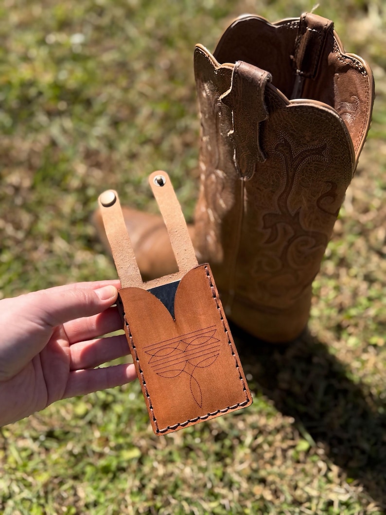 May include: A brown leather phone case with a decorative stitched design, held in front of a brown cowboy boot. The case has a flap with two rivets and a stitched border. The background is blurred green grass.
