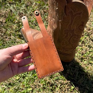 May include: A brown leather phone case with a strap and snap closure. The case is hand-stitched with a contrasting thread and is shown next to a brown leather cowboy boot. The case is designed to be attached to a belt or other item.