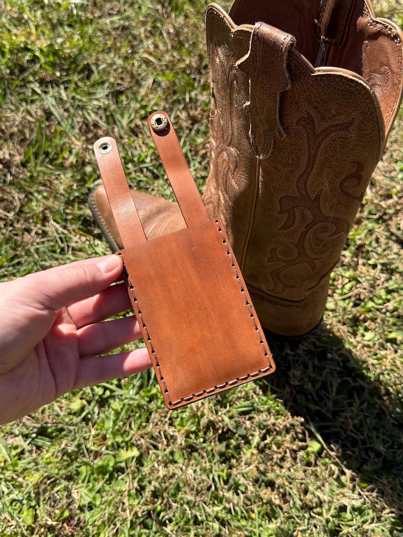 May include: A brown leather phone case with a strap and snap closure. The case is hand-stitched with a contrasting thread and is shown next to a brown leather cowboy boot. The case is designed to be attached to a belt or other item.