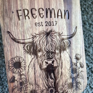 May include: Wooden cutting board with engraved design. Features a Highland cow illustration surrounded by sunflowers. The text "FREEMAN est 2017" is etched above the cow. The board has a natural wood grain finish.