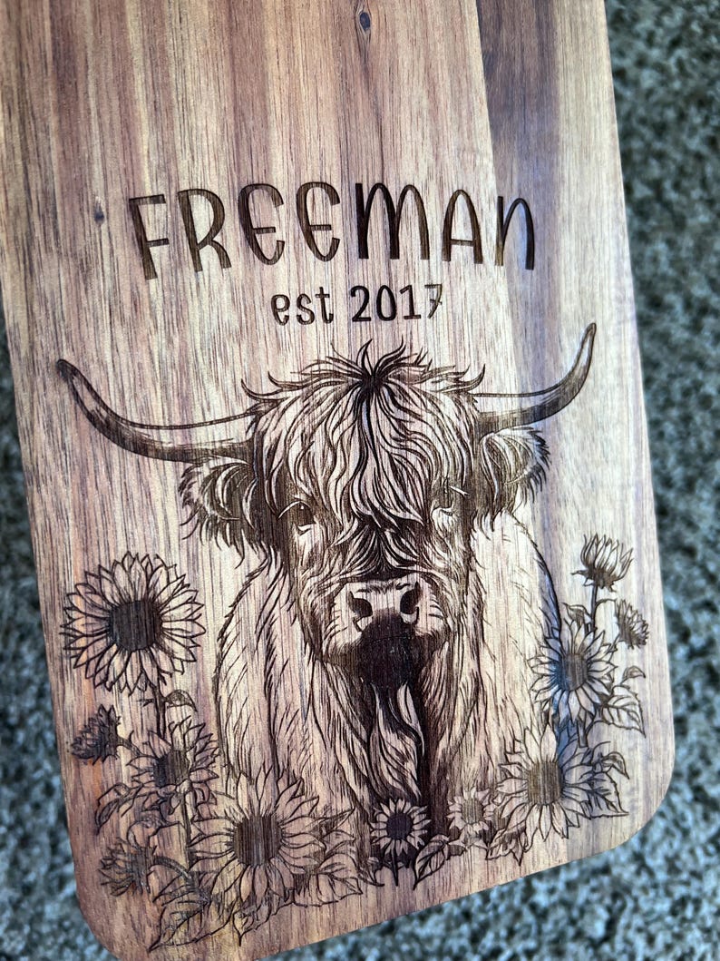 May include: Wooden cutting board with engraved design. Features a Highland cow illustration surrounded by sunflowers. The text "FREEMAN est 2017" is etched above the cow. The board has a natural wood grain finish.