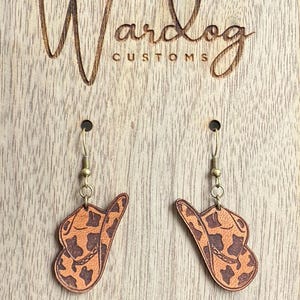 May include: Cowboy hat earrings with a brown and tan pattern, hanging from gold-colored hooks. The earrings are displayed on a wooden surface with the engraved text "Wardog Customs".