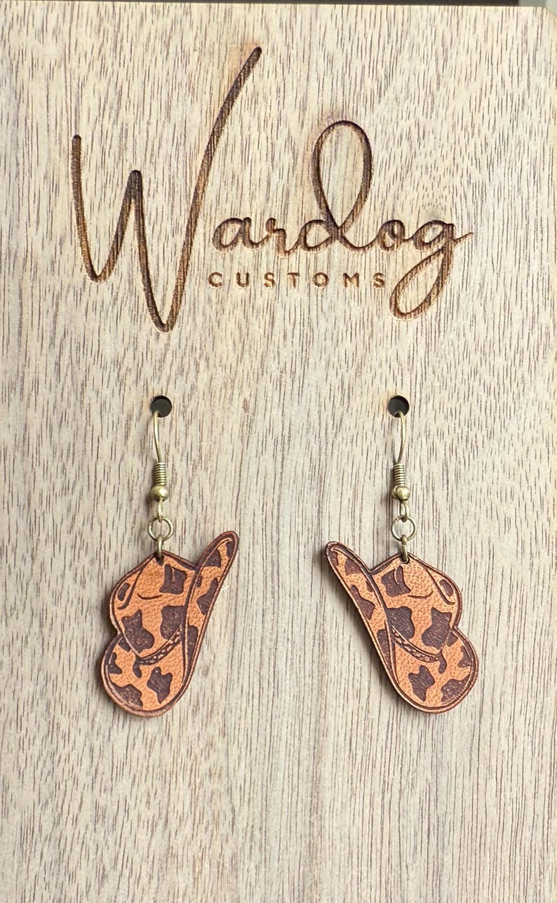 May include: Cowboy hat earrings with a brown and tan pattern, hanging from gold-colored hooks. The earrings are displayed on a wooden surface with the engraved text "Wardog Customs".