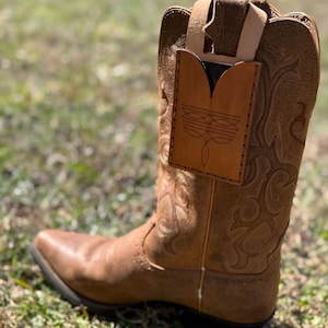 May include: A brown leather cowboy boot with decorative stitching and a pointed toe. A light brown leather phone holder is attached to the boot. The boot is set on a grassy surface.
