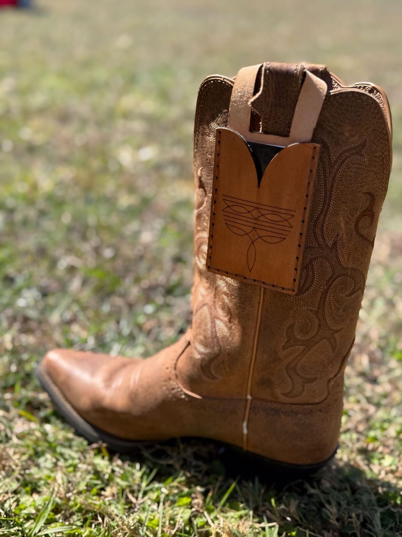 May include: A brown leather cowboy boot with decorative stitching and a pointed toe. A light brown leather phone holder is attached to the boot. The boot is set on a grassy surface.