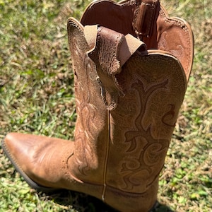 May include: A brown leather cowboy boot with intricate stitched floral patterns. The boot has a pointed toe and a worn appearance, suggesting regular use. The boot is set against a backdrop of green grass.