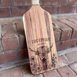 May include: Wooden cutting board with a handle wrapped in twine. The board features the name "FREEMAN" and "EST 2017" above an engraved image of a Highland cow surrounded by sunflowers. The board is made of wood.