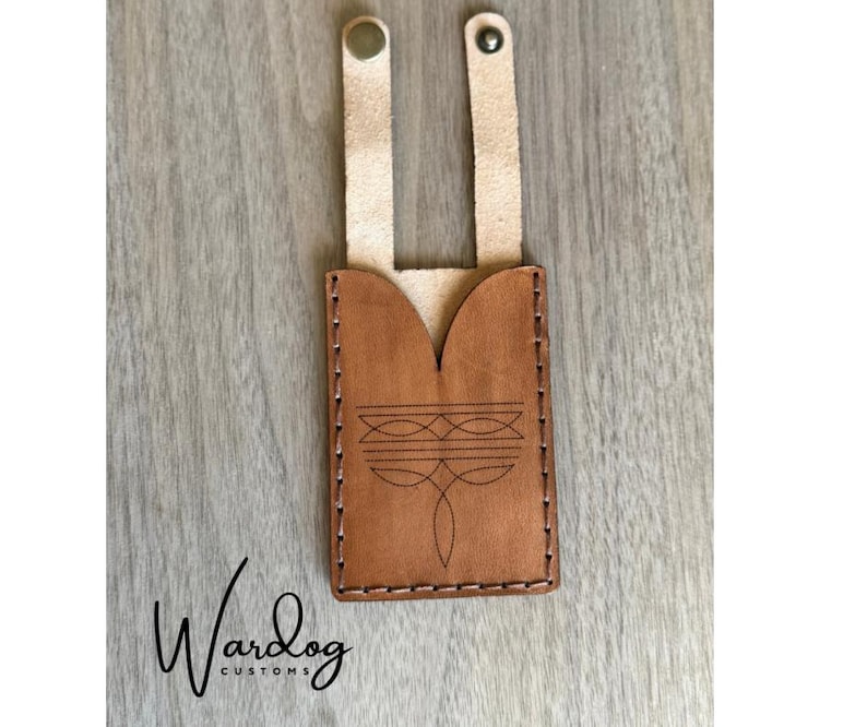 May include: A brown leather phone case with a decorative etched design. The case has two natural-colored leather straps with eyelets at the top. The brand name "Wardog Customs" is written in cursive at the bottom.