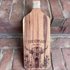 May include: Wooden cutting board with a handle and a decorative rope wrap. The board features the name "FREEMAN" and "est 2017" above an engraved image of a Highland cow and sunflowers.
