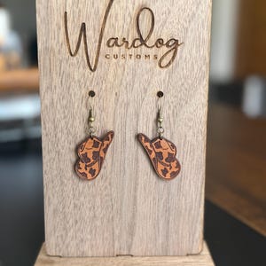 May include: A pair of earrings shaped like cowboy hats, featuring a brown and tan animal print design. The earrings hang from gold-colored hooks. The earrings are displayed on a wooden stand with the text "Wardog Customs".