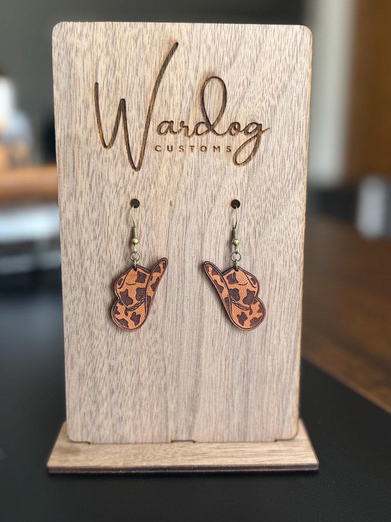 May include: A pair of earrings shaped like cowboy hats, featuring a brown and tan animal print design. The earrings hang from gold-colored hooks. The earrings are displayed on a wooden stand with the text "Wardog Customs".