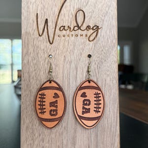 Op de afbeelding: Een paar leren oorbellen in de vorm van een voetbal met de letters "GA" en een hart, tentoongesteld op een houten standaard. De oorbellen zijn bruin met donkerbruine stikseldetails. De standaard heeft de tekst "Wardog Customs" gegraveerd.