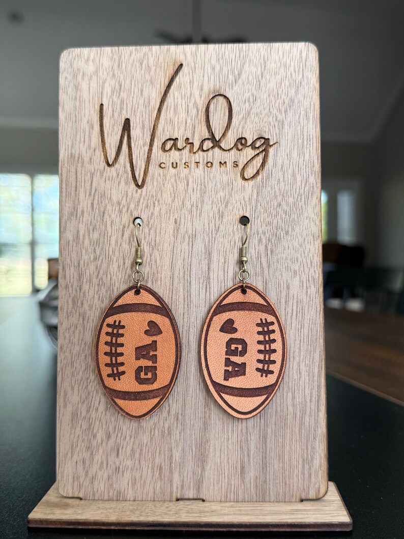 Op de afbeelding: Een paar leren oorbellen in de vorm van een voetbal met de letters "GA" en een hart, tentoongesteld op een houten standaard. De oorbellen zijn bruin met donkerbruine stikseldetails. De standaard heeft de tekst "Wardog Customs" gegraveerd.