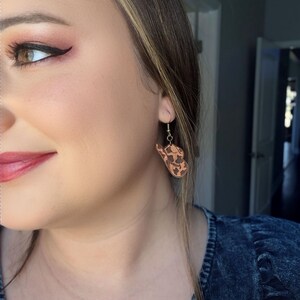 May include: Wooden earrings with a cowhide pattern are displayed. The earrings are hanging from a gold-colored hook. The earrings are a light brown color with dark brown spots. The earrings are worn by a person.