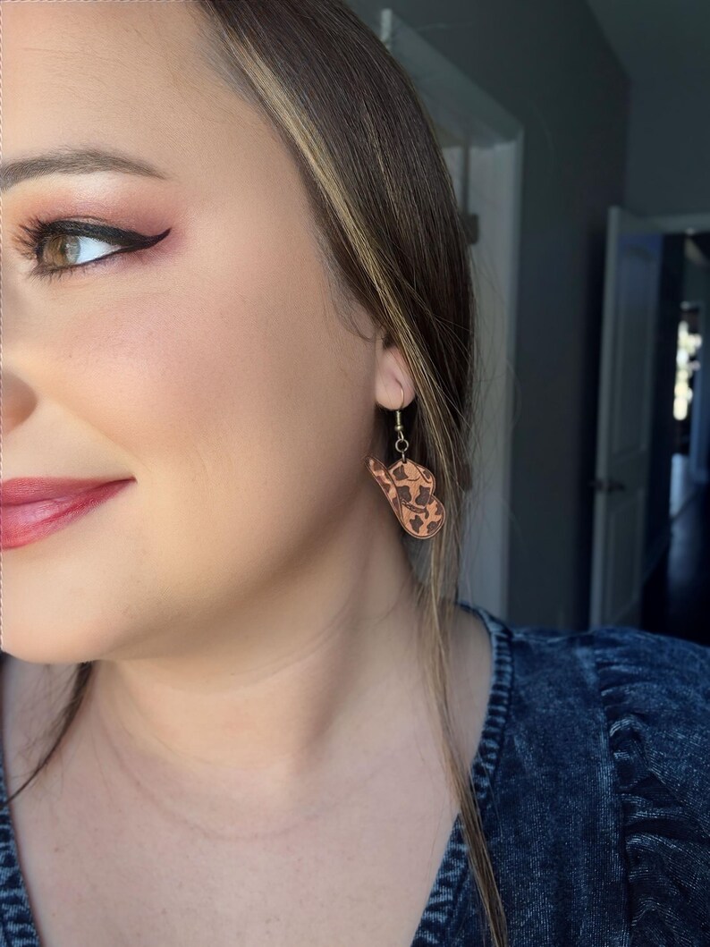 May include: Wooden earrings with a cowhide pattern are displayed. The earrings are hanging from a gold-colored hook. The earrings are a light brown color with dark brown spots. The earrings are worn by a person.