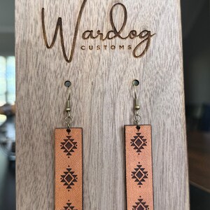 May include: Tan leather earrings with a geometric design are displayed on a wooden stand. The earrings are rectangular with a dark brown pattern. The wooden stand has the words "Wardog Customs" engraved on it.