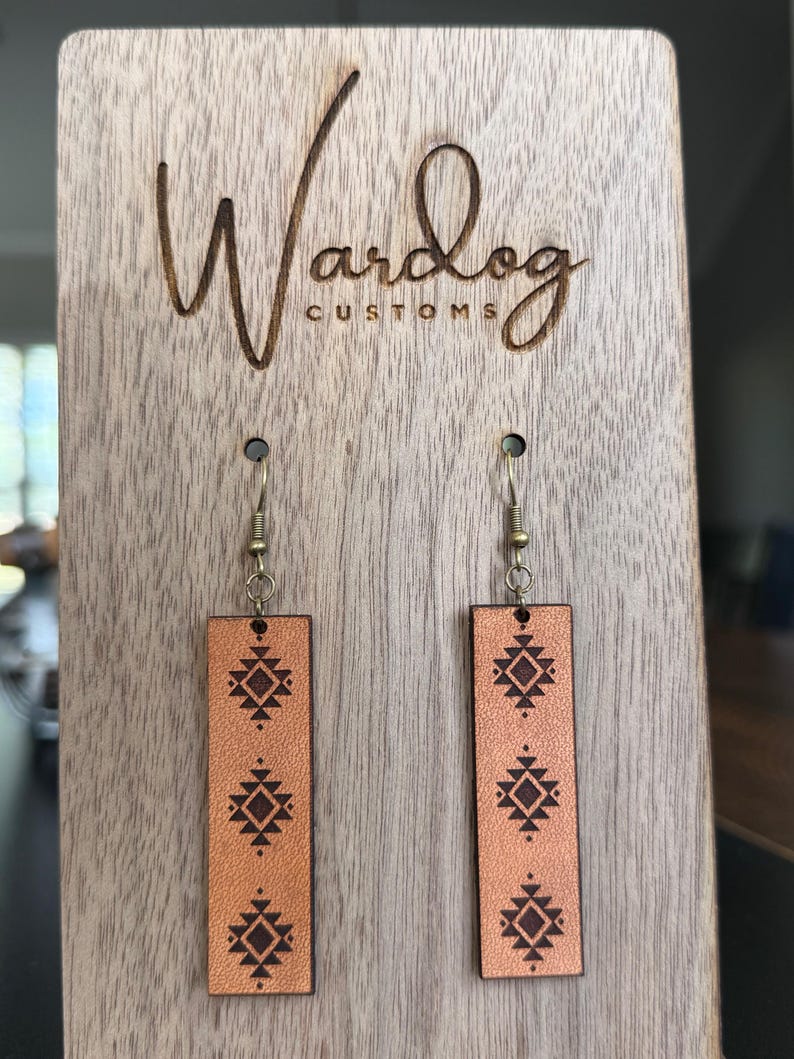 May include: Tan leather earrings with a geometric design are displayed on a wooden stand. The earrings are rectangular with a dark brown pattern. The wooden stand has the words "Wardog Customs" engraved on it.
