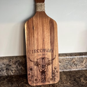 May include: Wooden cutting board with a handle wrapped in twine. The board features an engraved design of a Highland cow with the text "FREEMAN est 2017" and sunflowers. The board is made of a light brown wood.