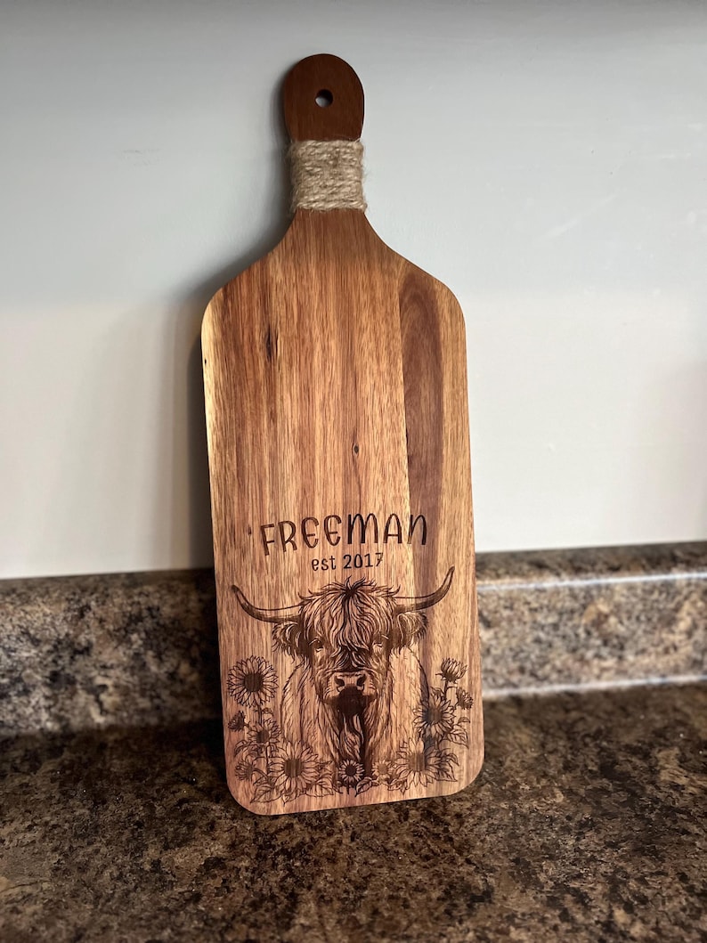 May include: Wooden cutting board with a handle wrapped in twine. The board features an engraved design of a Highland cow with the text "FREEMAN est 2017" and sunflowers. The board is made of a light brown wood.