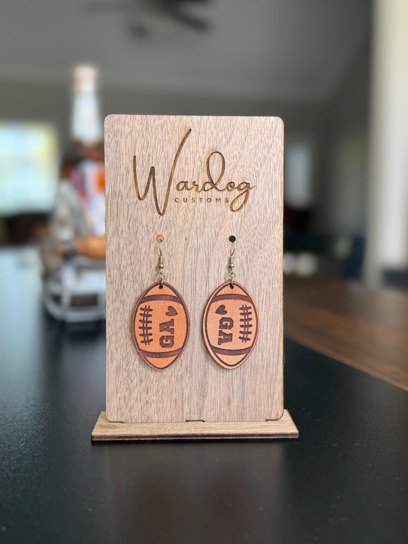 Op de afbeelding: Een paar oorbellen in de vorm van een voetbal met de letters "GA" en gestikte details. De oorbellen worden getoond op een houten standaard met het logo "Wardog Customs". De oorbellen zijn lichtbruin.