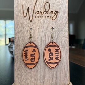 Op de afbeelding: Leren oorbellen in de vorm van een voetbal, met de letters "GA" en stikseldetails. Gepresenteerd op een houten standaard met de tekst "Wardog Customs".