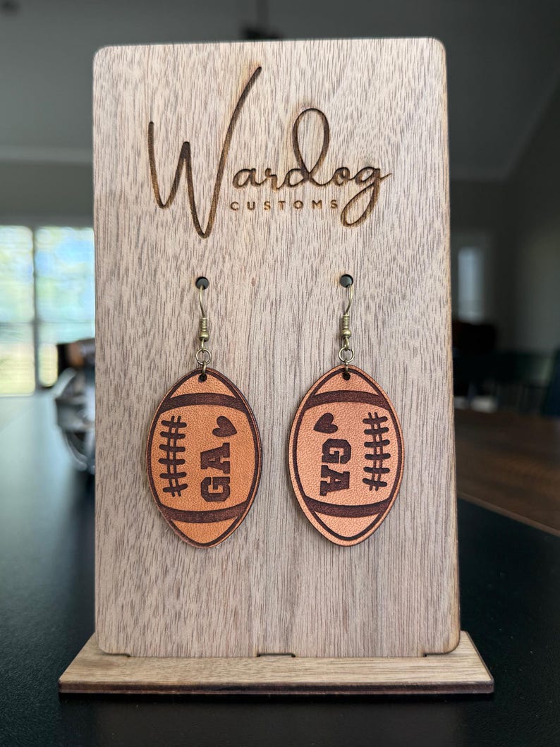 Op de afbeelding: Leren oorbellen in de vorm van een voetbal, met de letters "GA" en stikseldetails. Gepresenteerd op een houten standaard met de tekst "Wardog Customs".
