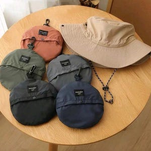 May include: A beige wide-brimmed sun hat with a black and white chin cord, accompanied by five circular, foldable storage pouches in terracotta, olive green, charcoal gray, black, and navy blue, each featuring a black label reading Packable Hat.