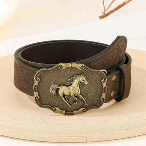 May include: A brown leather belt with an ornate, embossed design. The belt buckle features a gold-toned horse design.