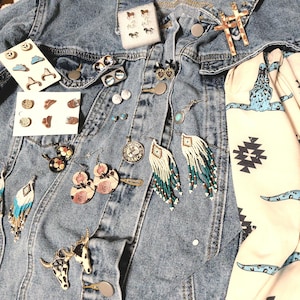 May include: A collection of Western-themed jewelry and accessories displayed on a denim jacket. Includes earrings with turquoise and white beading, floral earrings, pins, a scarf with a turquoise bull design, and a cross pendant.