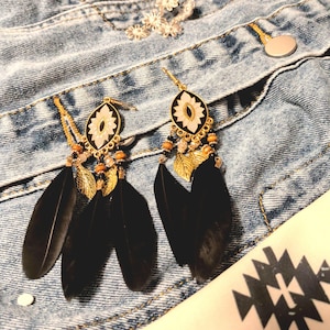 May include: A pair of dangling earrings with black feathers, gold-colored metal accents, and a floral design. The earrings are set against a denim jacket.