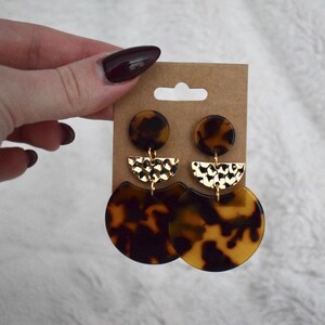 May include: A pair of gold and brown statement earrings with a tortoiseshell pattern. The earrings feature a large round disc with a smaller half-circle piece above it.