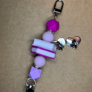 Puede incluir: Un llavero de cuentas de color morado y blanco con un dije de plata y dos dijes de libros apilados. El llavero tiene un broche de plata.
