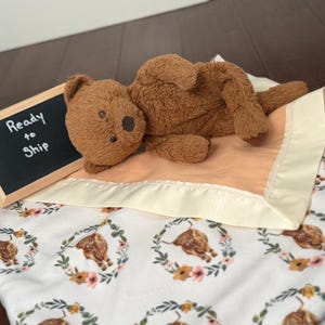 May include: A brown teddy bear rests on a peach-colored blanket with a cream satin border. The blanket features a white background with a floral and cow print. A small chalkboard sign reads "Ready to Ship."