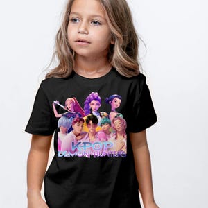 May include: Black t-shirt featuring a colorful graphic of a group of animated characters with the text "KPOP DEMON HUNTERS" in a stylized font. The design includes various hair colors and styles. The shirt is worn by a young person.