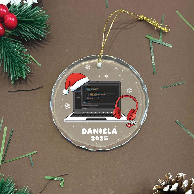 K&ouml;nnte beinhalten: Ein klarer, runder Weihnachtsschmuck mit einem goldenen Faden. Der Schmuck zeigt einen Laptop mit Code, einen Weihnachtsmannhut und rote Kopfh&ouml;rer. Der Name "DANIELA" und das Jahr "2025" sind aufgedruckt.
