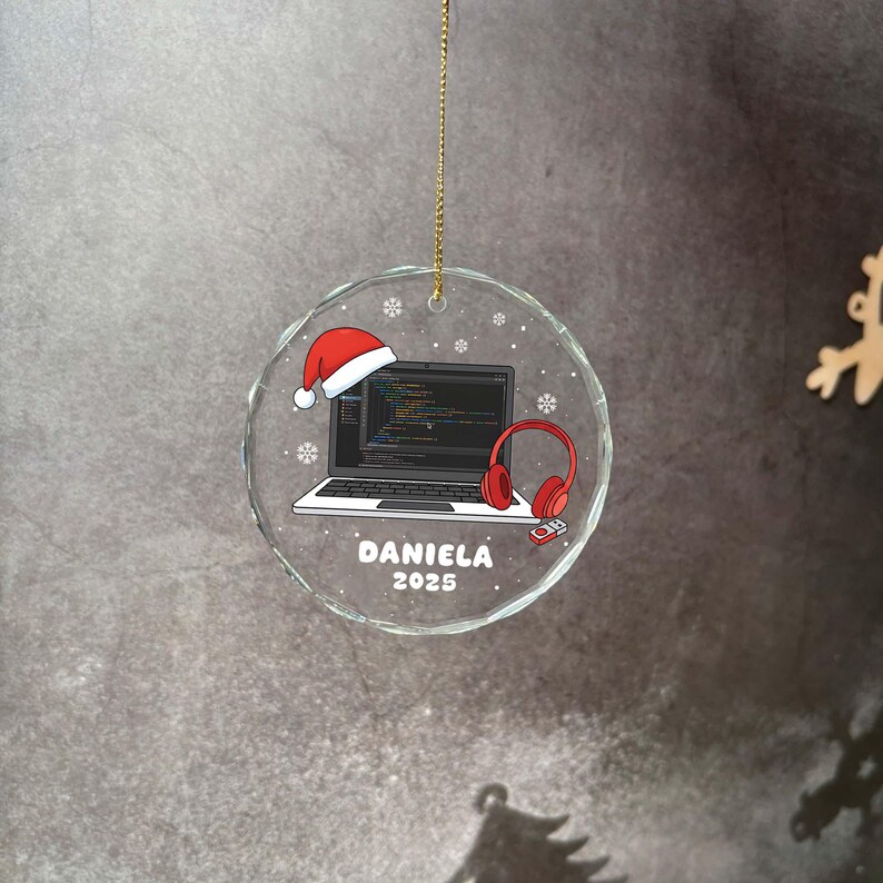 K&ouml;nnte beinhalten: Transparenter Weihnachtsbaumschmuck aus Glas mit goldenem Faden. Der Schmuck zeigt einen Laptop mit Code auf dem Bildschirm, einen Weihnachtsmannhut und rote Kopfh&ouml;rer. Der Name "DANIELA" und das Jahr "2025" sind aufgedruckt.