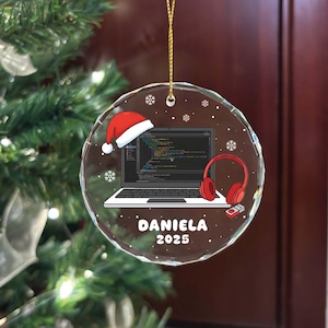 Puede incluir: Adorno navideño redondo y transparente con una cuerda dorada. Presenta un portátil con código, un gorro de Papá Noel, auriculares rojos y el nombre "DANIELA 2025". Copos de nieve decoran el diseño.