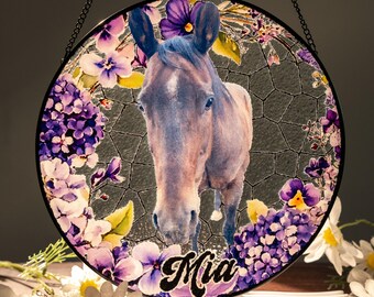 Pode incluir: Painel redondo de vitral com um retrato de cavalo rodeado por flores roxas e brancas. O cavalo é castanho com uma crina azul e o nome "Mia" está escrito em itálico na parte inferior. O painel é pendurado por uma corrente preta.