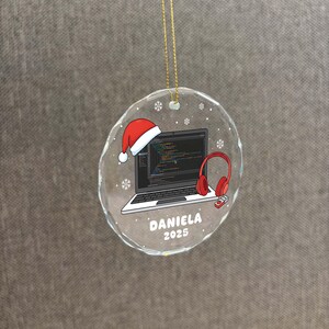 K&ouml;nnte beinhalten: Runder, klarer Weihnachtsschmuck mit goldenem Faden. Der Schmuck zeigt einen Laptop mit Code auf dem Bildschirm, einen Weihnachtsmannhut, rote Kopfh&ouml;rer und eine Fernbedienung. Der Name "DANIELA" und das Jahr "2025" sind aufgedruckt.