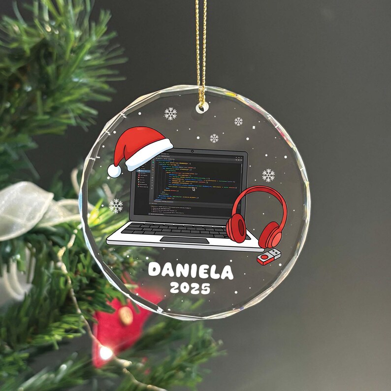 K&ouml;nnte beinhalten: Ein klarer, runder Weihnachtsschmuck mit einem goldenen Faden. Der Schmuck zeigt einen Laptop mit Code, einen Weihnachtsmannhut, rote Kopfh&ouml;rer und den Namen "Daniela" mit dem Jahr "2025". Schneeflocken sind um das Design verstreut.