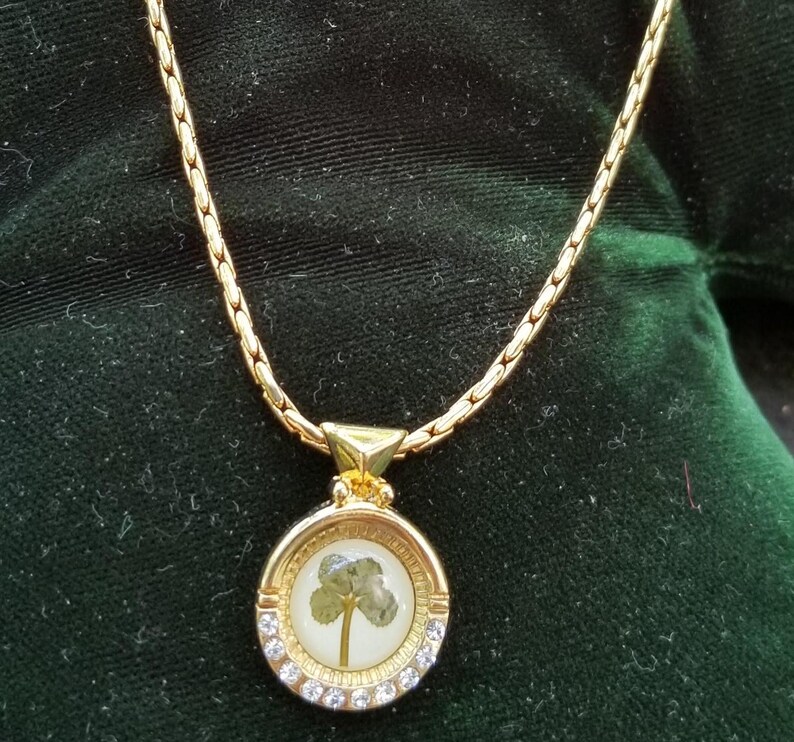 Peut inclure: Collier dor&eacute; avec un pendentif rond orn&eacute; d'un tr&egrave;fle &agrave; quatre feuilles encapsul&eacute; dans de la r&eacute;sine transparente. Le pendentif est entour&eacute; de petits cristaux transparents. La cha&icirc;ne est textur&eacute;e.