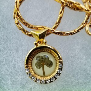 Peut inclure: Collier dor&eacute; avec un pendentif rond contenant un tr&egrave;fle &agrave; quatre feuilles encapsul&eacute; dans de la r&eacute;sine transparente. Le pendentif est orn&eacute; de petits cristaux transparents. La cha&icirc;ne est de conception d&eacute;licate et maill&eacute;e.