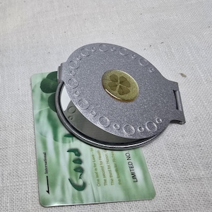 Op de afbeelding: Een zilveren compacte spiegel met een klavertjevier ingekapseld in hars. De spiegel is open en toont het reflecterende oppervlak. Een groene kaart met het woord "Good" en tekst over de betekenis van de klaver is gedeeltelijk zichtbaar.