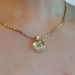 Peut inclure: Collier dor&eacute; avec un pendentif en forme de tr&egrave;fle. Le pendentif rond pr&eacute;sente un fond blanc et un tr&egrave;fle &agrave; quatre feuilles vert. Le pendentif est entour&eacute; de petits cristaux transparents. La cha&icirc;ne est de conception d&eacute;licate et textur&eacute;e.
