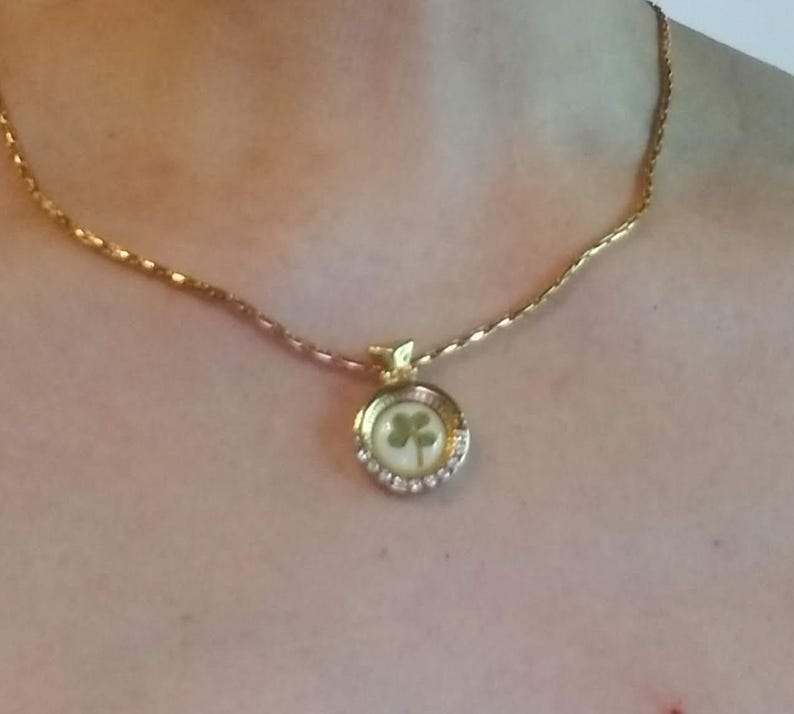 Peut inclure: Collier dor&eacute; avec un pendentif en forme de tr&egrave;fle. Le pendentif rond pr&eacute;sente un fond blanc et un tr&egrave;fle &agrave; quatre feuilles vert. Le pendentif est entour&eacute; de petits cristaux transparents. La cha&icirc;ne est de conception d&eacute;licate et textur&eacute;e.
