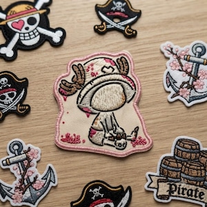 Könnte beinhalten: Eine Sammlung bestickter Aufnäher mit einer zentralen Figur, die einen Hut mit Geweih trägt, umgeben von Piratenmotiven wie Totenköpfen, Ankern mit rosa Kirschblüten und einem Stapel Fässer mit der Aufschrift Pirate.