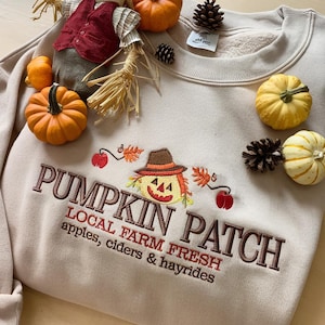 Könnte beinhalten: Beiges Sweatshirt mit dem gestickten Text "PUMPKIN PATCH" und einer Vogelscheuchen-Grafik. Das Sweatshirt ist von kleinen Kürbissen, Tannenzapfen und einer Vogelscheuchen-Figur umgeben. Der Text darunter lautet "LOCAL FARM FRESH apples, ciders & hayrides."