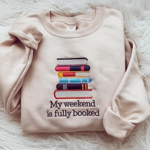 May include: A beige sweatshirt with a stack of embroidered books and the text "My weekend is fully booked".