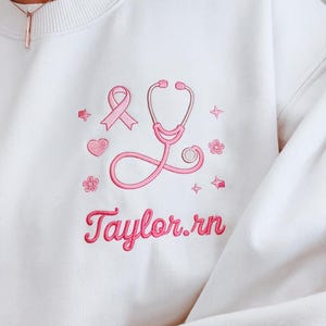 Könnte beinhalten: Cremefarbenes Sweatshirt mit rosa Stickerei. Es zeigt ein Stethoskop, ein Band, ein Herz, Sterne und Blumen. Der Name "Taylor.rn" ist ebenfalls rosa gestickt. Eine Halskette ist oben sichtbar.